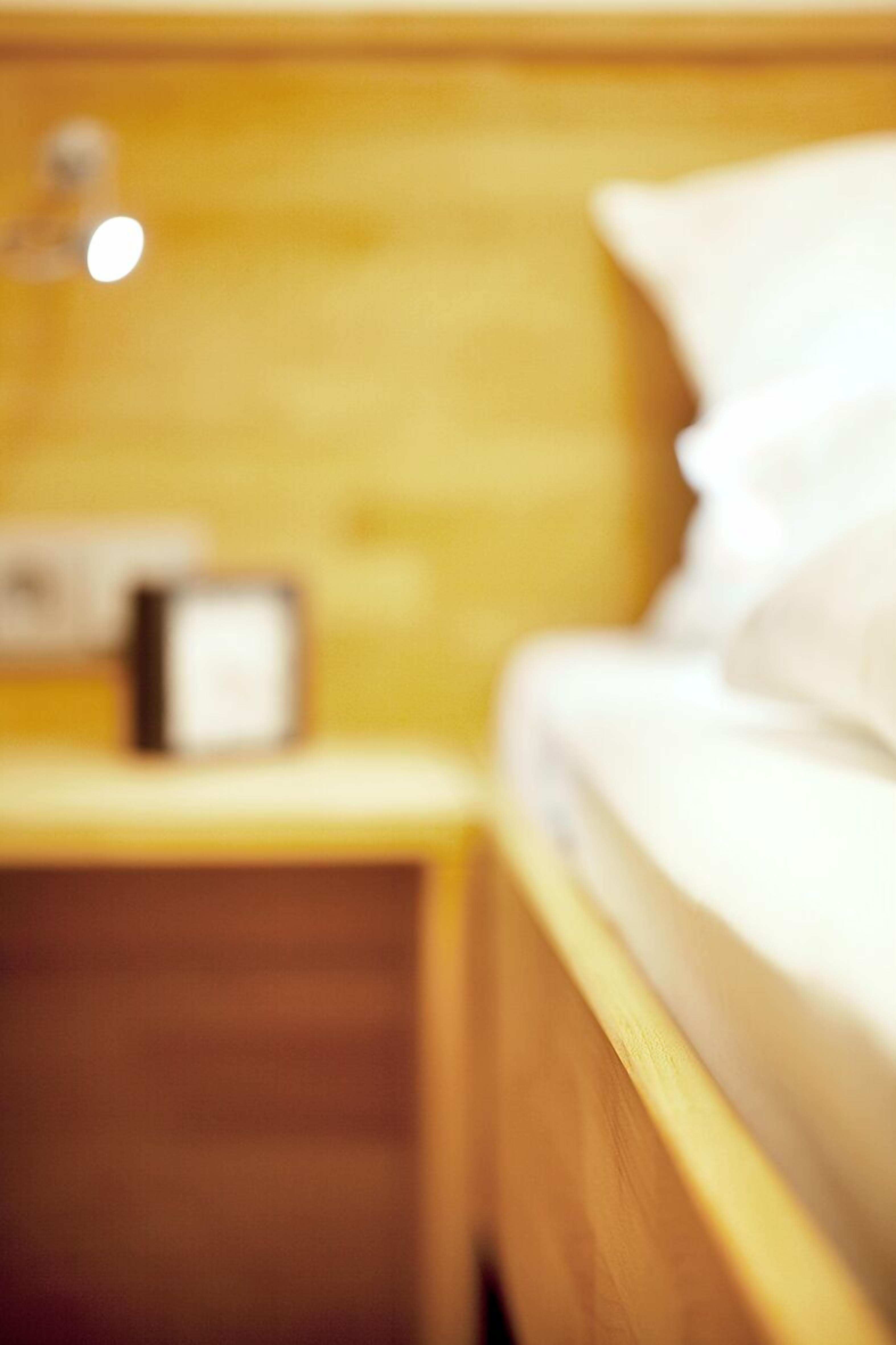 A blurred view of a wooden bed with white bedding and a nightstand with a lamp.