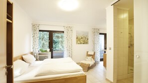 A bedroom with a wooden bed, a view of a forest through a glass door, and a bathroom with a glass shower. - Flörsbacher Hof (Floersbachtal)
