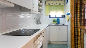 A compact kitchen with white cabinets, a black induction cooktop, a dishwasher, and a window with a yellow valance. - ApartUP Saplaya Harbour (Alboraya)