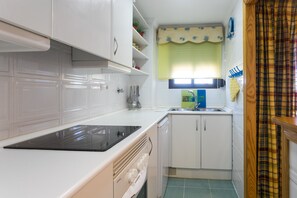 A compact kitchen with white cabinets, a black induction cooktop, a dishwasher, and a window with a yellow valance. - ApartUP Saplaya Harbour (Alboraya)