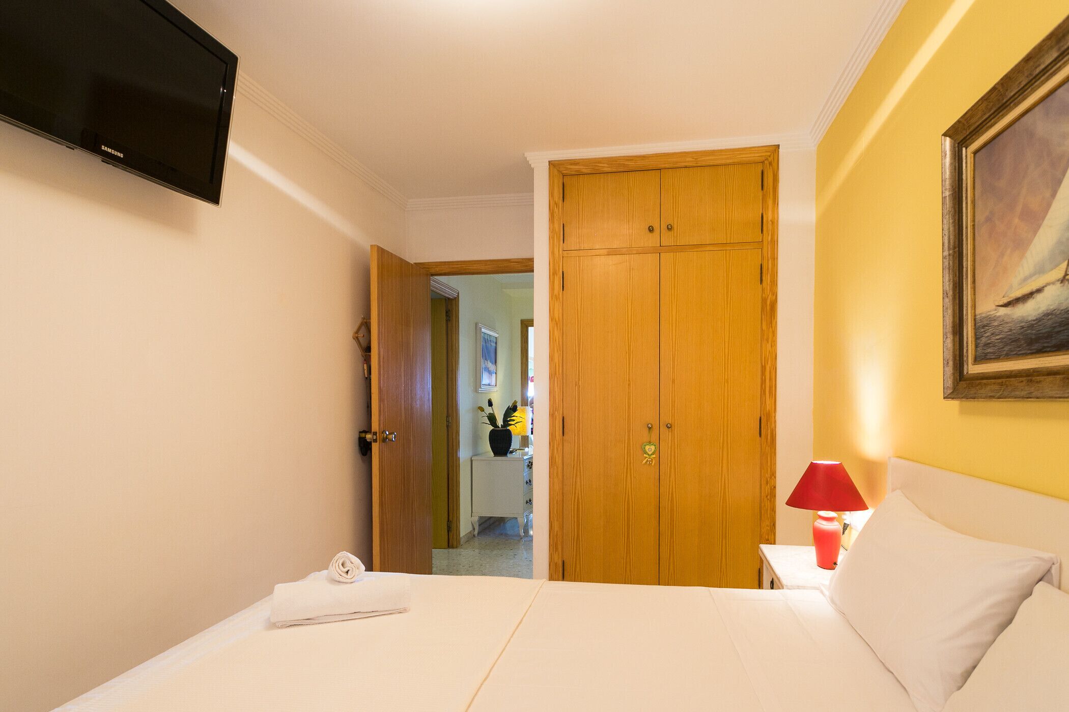 Une chambre d’hôtel avec un lit, un téléviseur fixé au mur, une armoire en bois et un tableau accroché au mur.