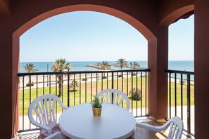 A balcony with a white table and chairs, overlooking a beach with palm trees and a clear sky.
