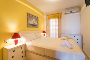 A neatly made bed with white linens, a red lamp on the nightstand, a painting of a sailboat on the wall, and a window with curtains.
