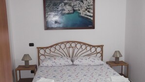 Una cama bien hecha con un edredón estampado, dos lámparas de noche y un cuadro enmarcado con una escena costera sobre la cama.