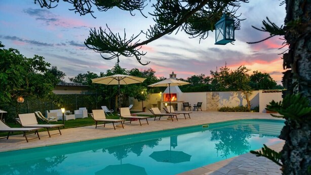 Una piscina con sillones de descanso y sombrillas, rodeada de follaje y un cielo al atardecer.