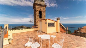 Una terraza con sillas y mesa de metal blanco, con vistas al mar, una torre de piedra y cielo despejado.