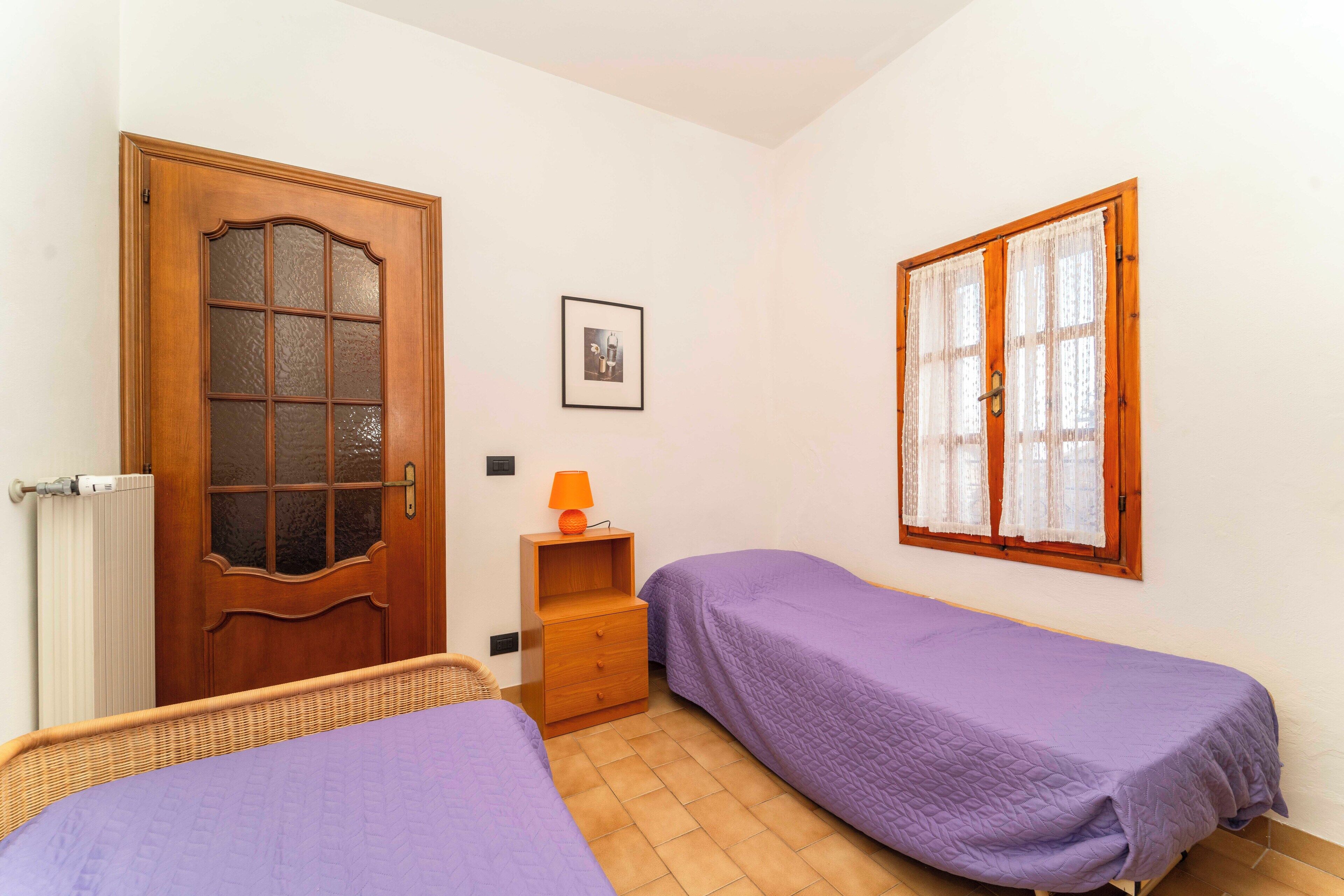 Un dormitorio con dos camas individuales, una puerta de madera, un ventanal con cortinas, una mesita de noche pequeña con una lámpara y un cuadro en la pared.