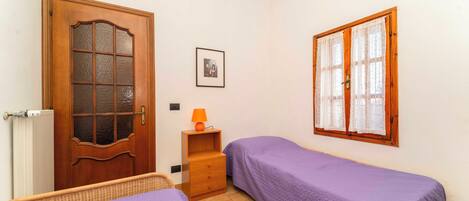Un dormitorio con dos camas individuales, una puerta de madera, un ventanal con cortinas, una mesita de noche pequeña con una lámpara y un cuadro en la pared.