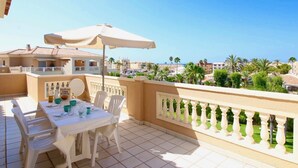 A balcony with a table set for dining, a white umbrella, and a view of the sea and buildings.