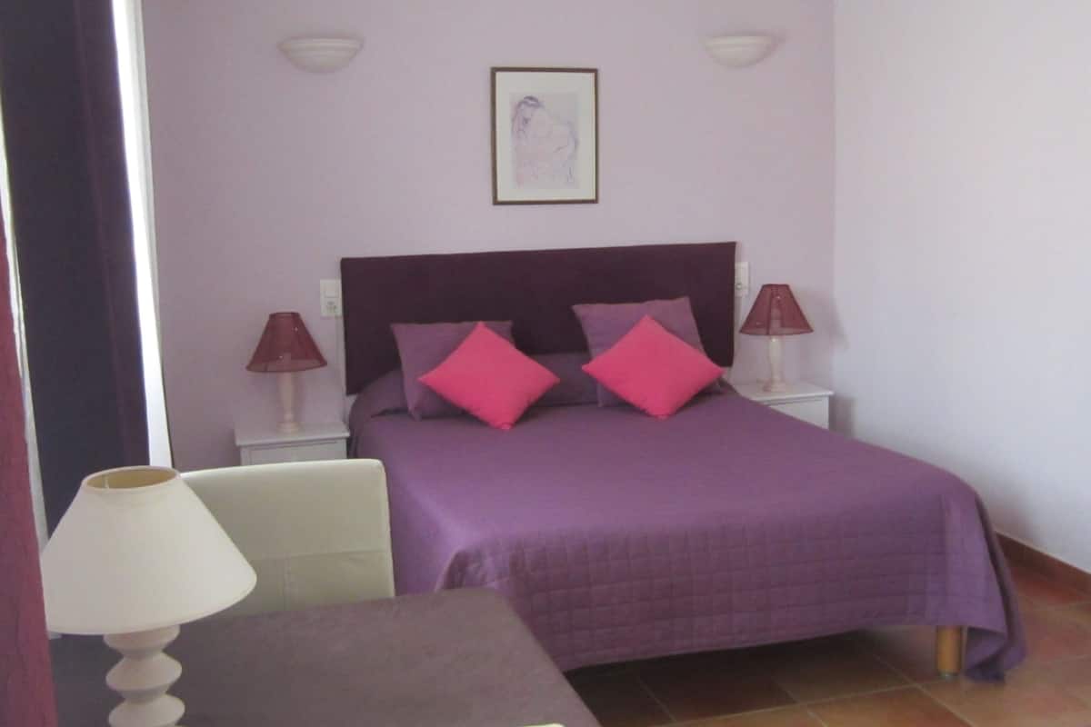 A bedroom with a bed, two bedside lamps, a framed picture on the wall, and a chair with a table.