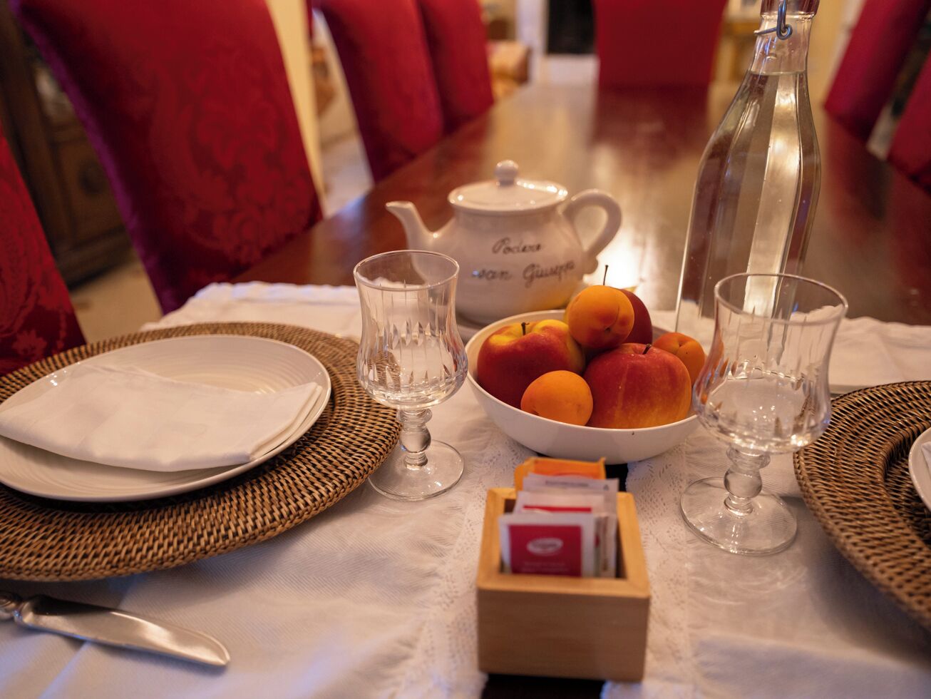 A dining table set with plates, glasses, a teapot, a bottle, and a bowl of fruit.