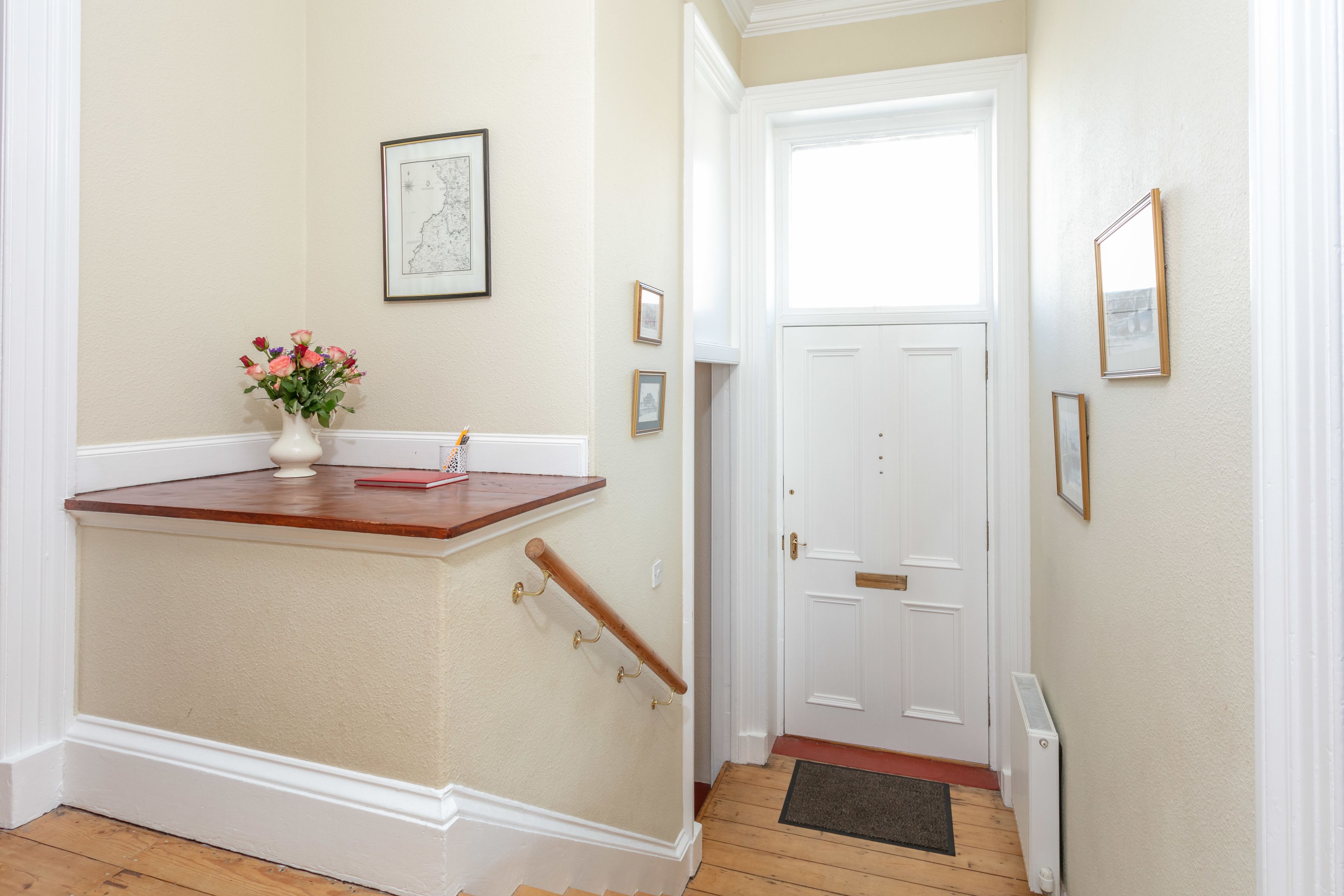 Um corredor com corrimão de madeira, uma porta branca e um pequeno vaso com flores sobre um pedestal branco.