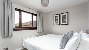 A bedroom with a large bed, grey curtains, a window, and two framed pictures on the wall. - The Huntington - Donnini Apartments (Ayr)