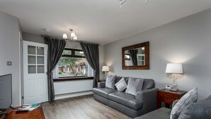 A modern living room with a grey sofa, a wooden coffee table, and a TV. There is a window with curtains, a mirror on the wall, and a lamp on a side table. - The Huntington - Donnini Apartments (Ayr)