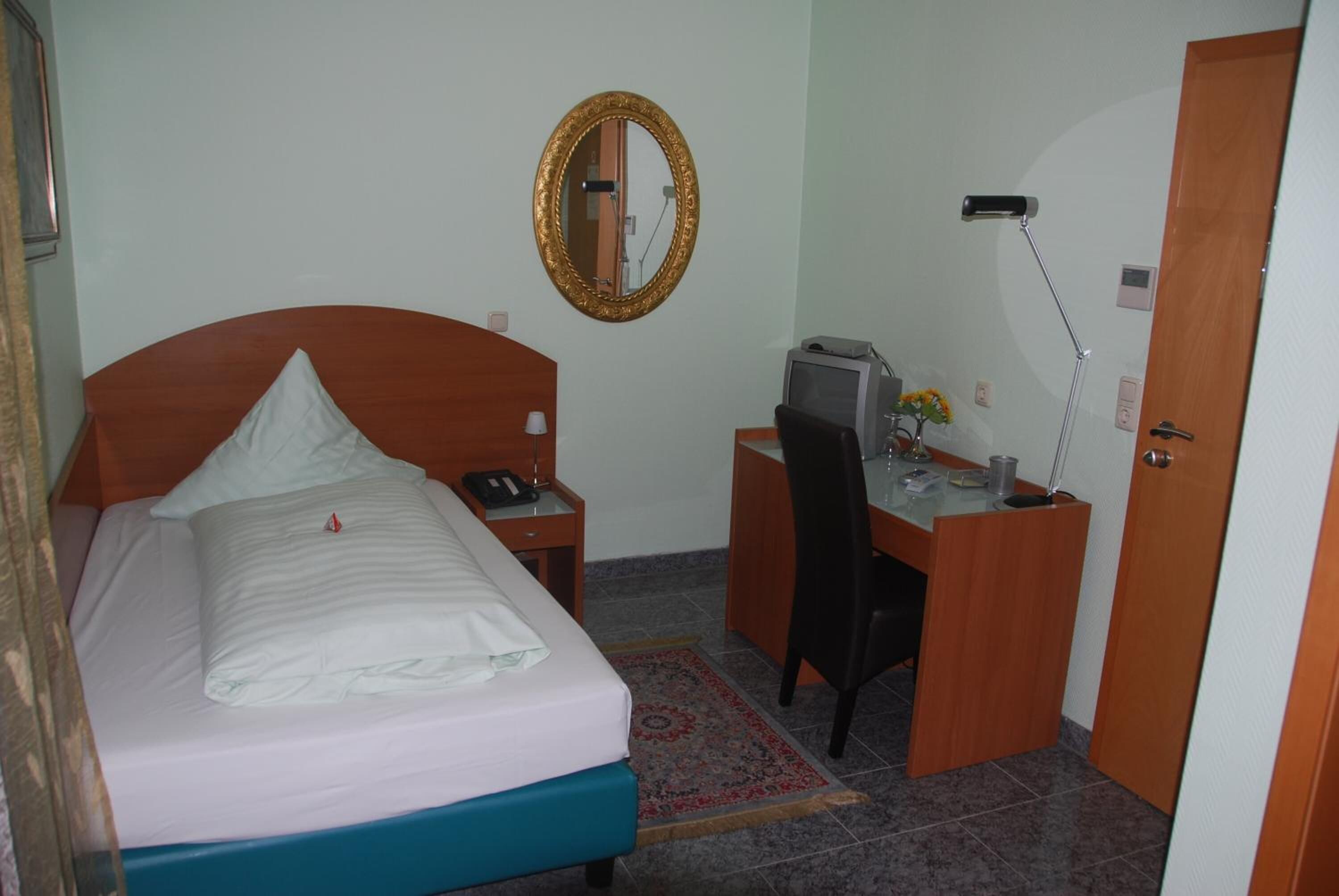 A hotel room with a bed, a desk, a chair, a telephone, a mirror, and a door.