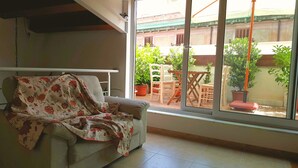 A living room with a sofa covered in a floral blanket, a window with a view of a courtyard with potted plants and outdoor furniture. - PalermiT'Amo Apartment (Palermo)