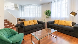 Una sala de estar moderna con dos sofás oscuros, una mesa de centro de vidrio y un sillón verde. Se ve una escalera al fondo.