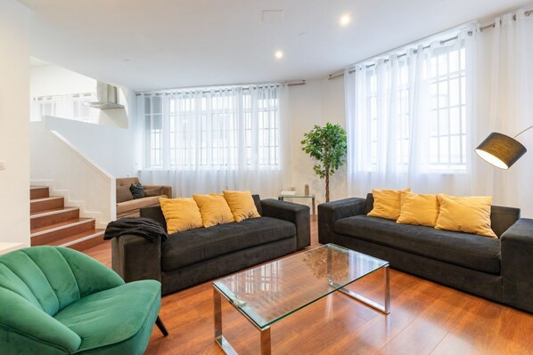 A modern living room with two dark sofas, a glass coffee table, and a green armchair. A staircase is visible in the background.