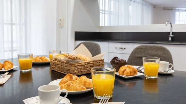 Un desayuno con croissants, zumo de naranja y café sobre una mesa en una cocina moderna.