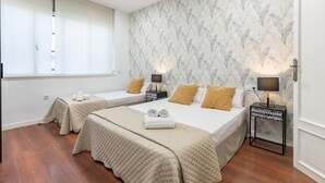 Habitación de hotel con dos camas, un ventanal grande con persianas, un papel tapiz estampado y una mesita con lámpara.