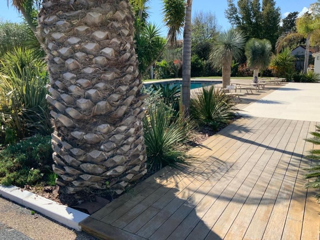 Una terraza de madera con un pilar de piedra, rodeada de plantas diversas y una piscina al fondo.