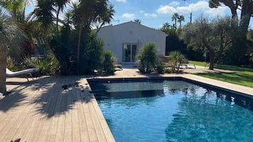 A swimming pool with a wooden deck, surrounded by palm trees and a building with a white facade.
