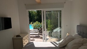 A bedroom with a bed, a TV, a small bedside table, and a sliding glass door leading to an outdoor area with a pool and lounge chairs. - Guesthouse Le Mas de la Dame (Saint-Tropez)