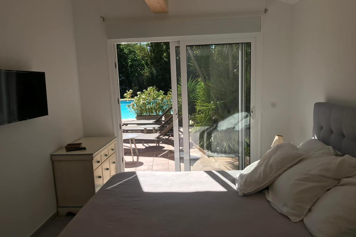 A bedroom with a bed, a TV, a small bedside table, and a sliding glass door leading to an outdoor area with a pool and lounge chairs.