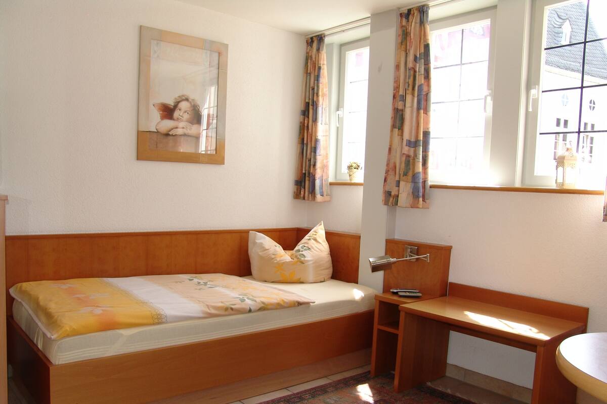 Een knus, klein kamertje met een enkel bed, een bureau en een stoel. Er is een raam met gordijnen, een klein tafeltje en een wandplank.