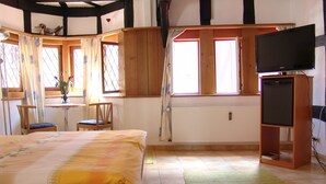 Ein Schlafzimmer mit Bett, Sessel, kleinem Tisch, Fernseher auf einem Schrank und einem Fenster mit Vorhängen.