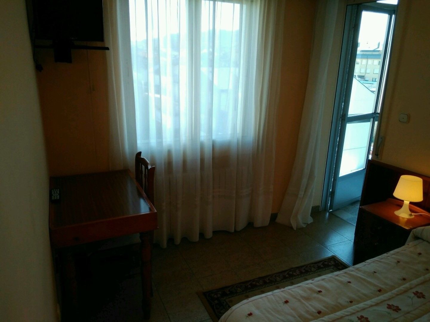 Una habitación con cama, mesa, silla, lámpara y ventana con cortinas.