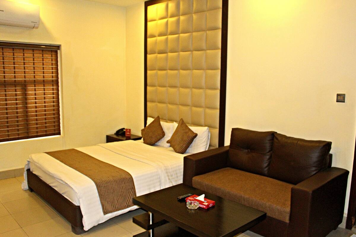 A hotel room with a bed, a brown leather sofa, a small table, and a window with blinds.