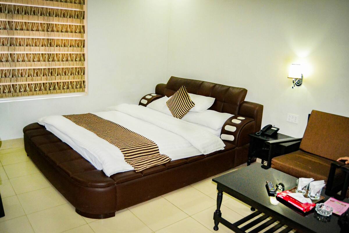A neatly made bed with a striped blanket, a brown leather sofa, a small table with a remote control, and a wall-mounted lamp.