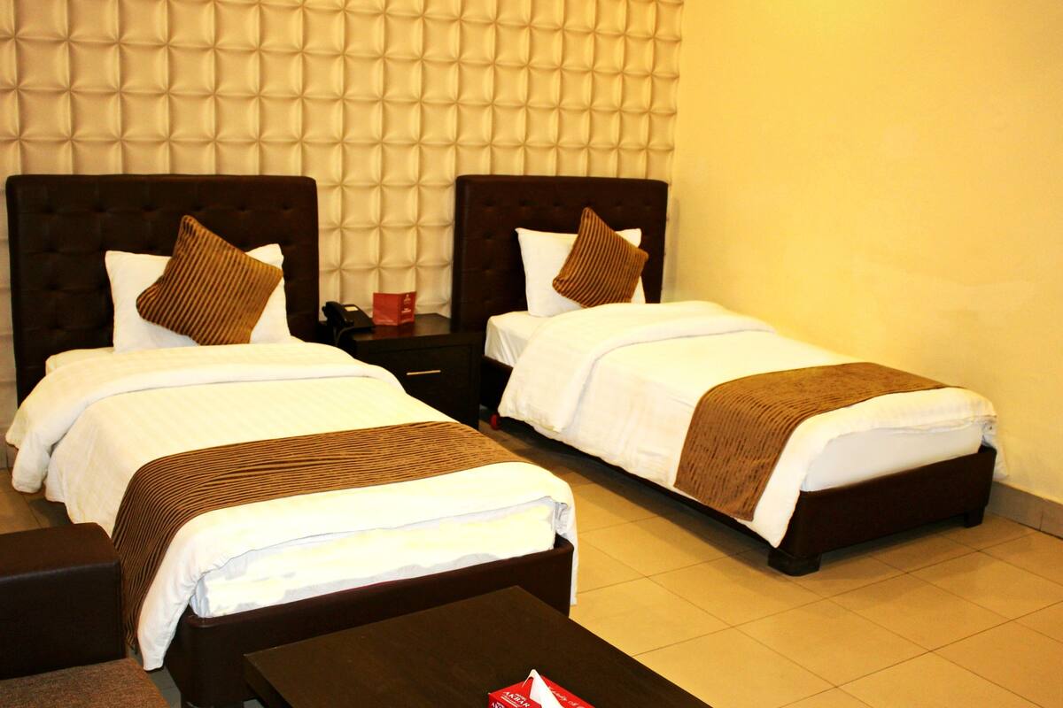 A hotel room with two beds, each with a brown headboard, white bedding, and a brown throw blanket. There is a small table between the beds and a red object on the nightstand.