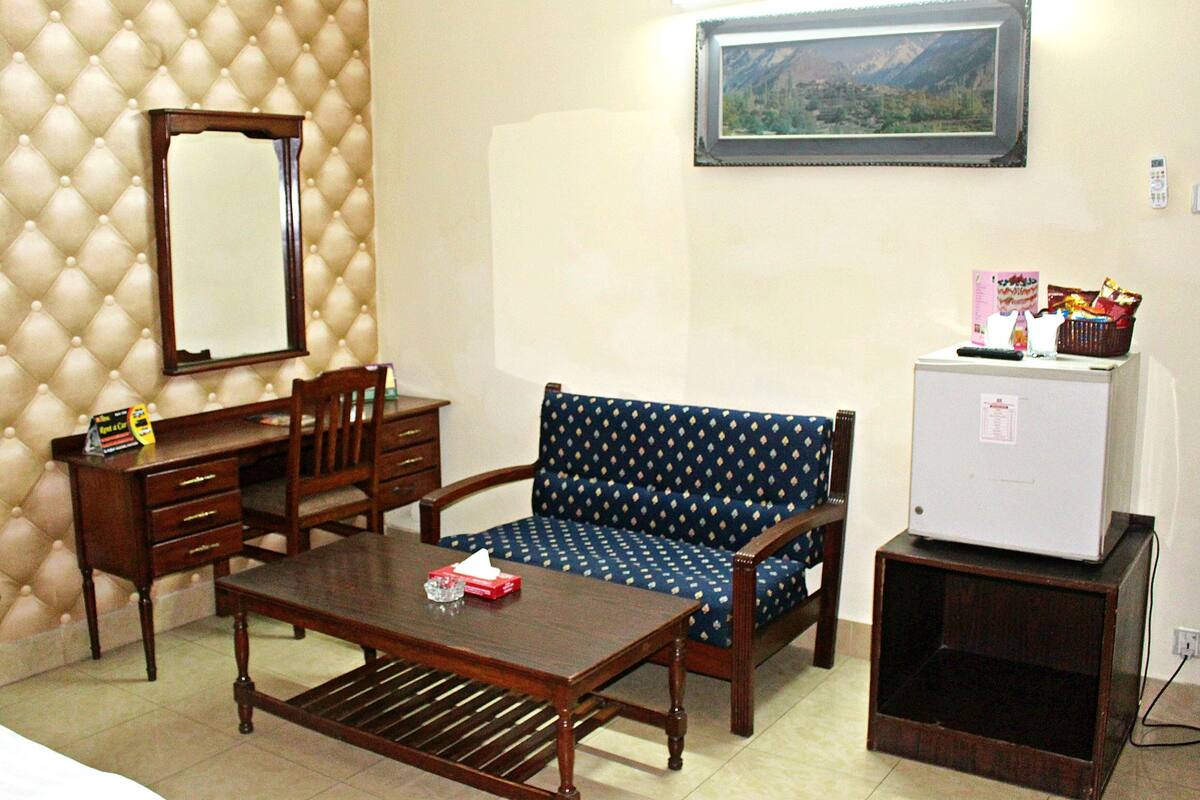 A room with a tufted wall, a wooden desk with drawers, a patterned sofa, a small refrigerator, and a framed picture on the wall.