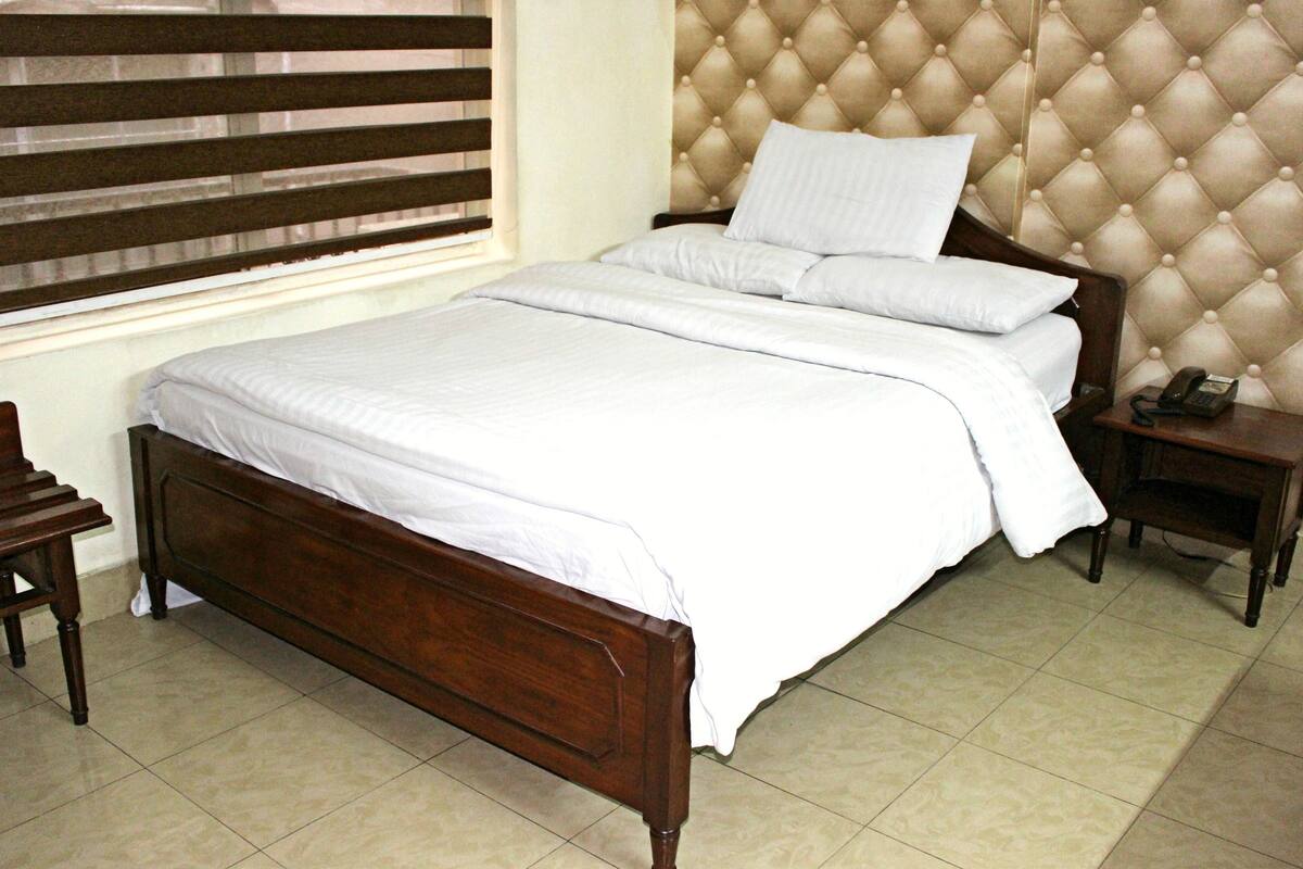 A neatly made bed with a wooden frame and white bedding, a wooden nightstand with a telephone, and a window with blinds.