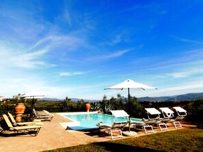 A pool area with lounge chairs and umbrellas, surrounded by a clear sky and distant mountains.