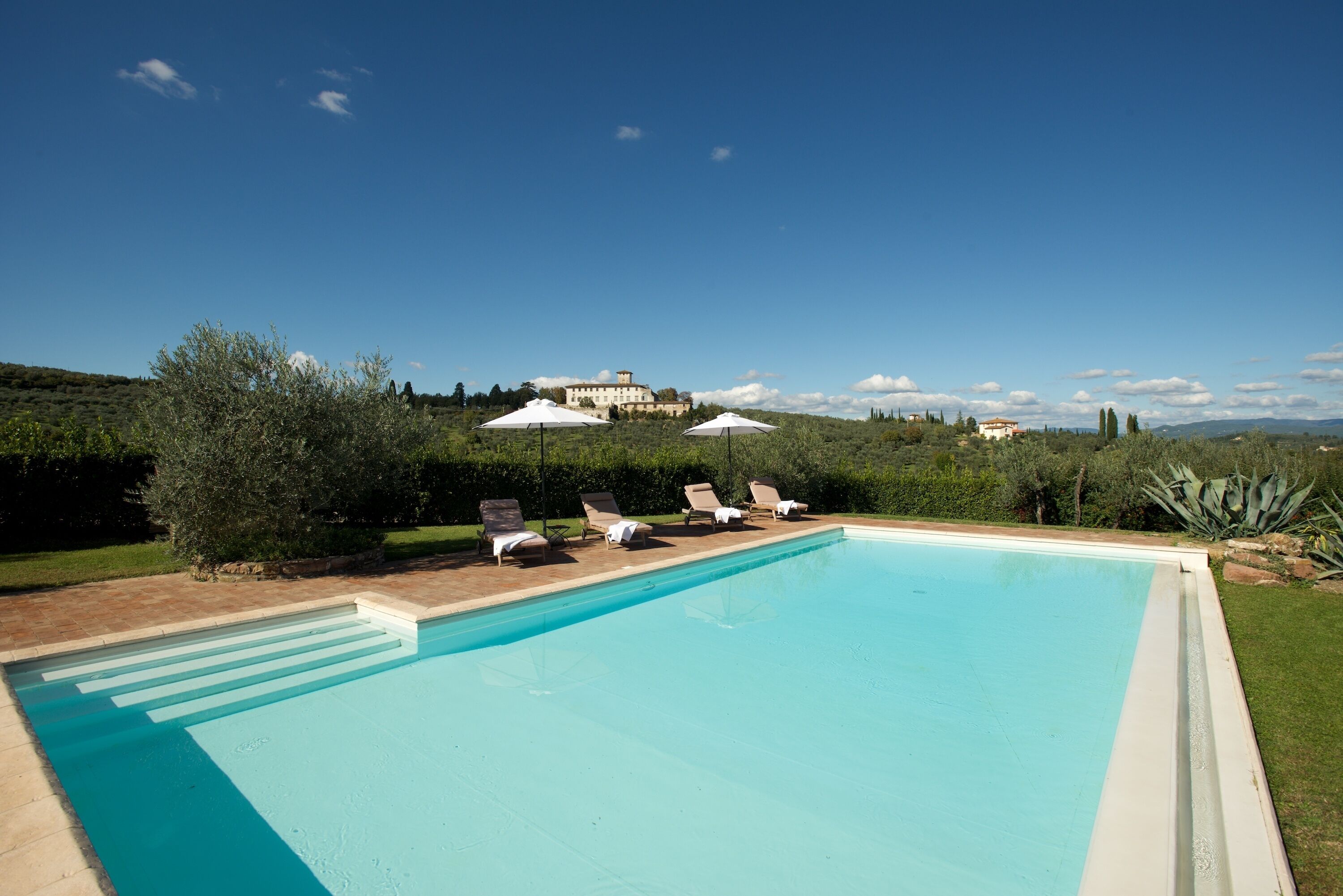 Une piscine entourée de chaises longues et de parasols, bordée de verdure et offrant une vue lointaine sur une colline où se trouve un bâtiment.