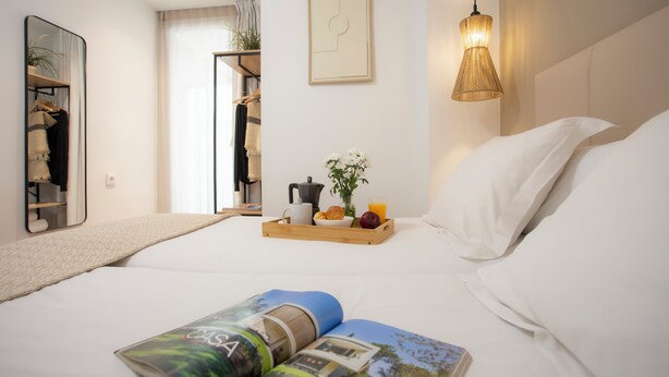 A hotel room with a bed, a bedside table with a tray of breakfast, a hanging light fixture, a mirror, and a framed picture on the wall. - Singular Stays Mercado Central VII (Valencia ciudad)