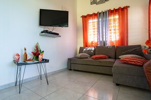 A living room with a flat-screen TV mounted on the wall, a sofa with cushions, a small table with a bottle and decorative items, and a window with orange curtains.