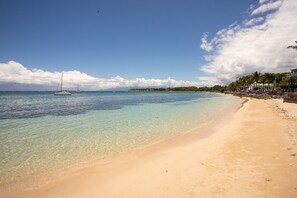 A beach with clear turquoise water, a sandy shore, and a sailboat in the distance.
