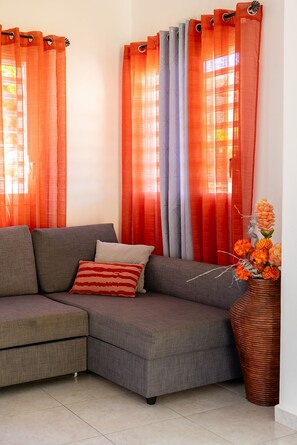 Un salón con un sofá seccional gris, un cojín a rayas rojo y blanco, y un jarrón con flores naranjas.