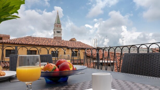 Una mesa de desayuno con un vaso de jugo de naranja, un bol de fruta y una taza blanca en una terraza con vistas a un edificio con una torre.