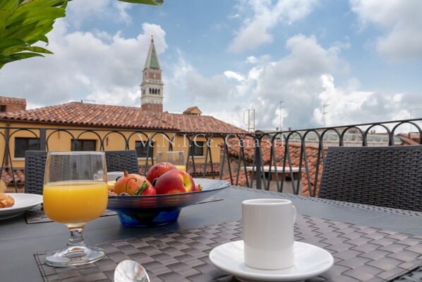 A breakfast table with a glass of orange juice, a bowl of fruit, and a white cup on a terrace with a view of a building with a tower.