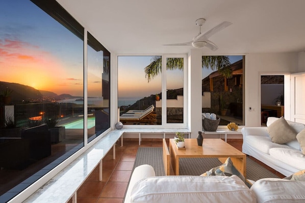 A modern living room with large windows, a ceiling fan, and a view of a pool and mountains.