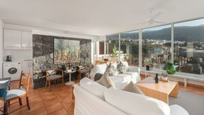 A modern living room with a stone wall, white furniture, and a large window offering a city view. - Plunge pool, terraces, garden, BBQ & lounge [A] (Icod de los Vinos)
