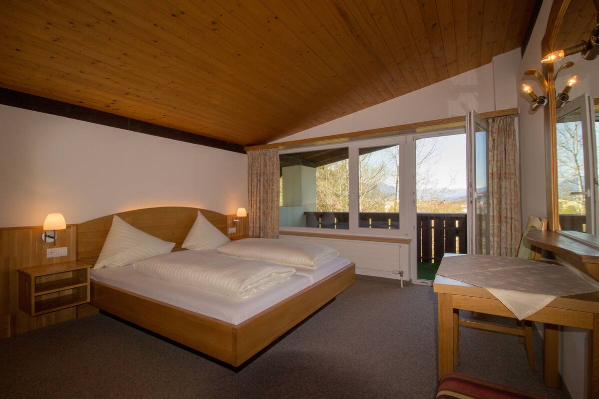 A bedroom with a wooden ceiling, a large bed with white linens, a nightstand with a lamp, a window with curtains, and a view of the outdoors.