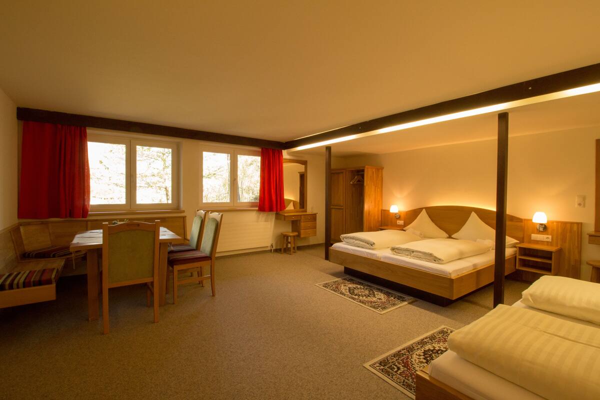 A hotel room with a bed, a desk with a chair, a small table, and a window with red curtains.