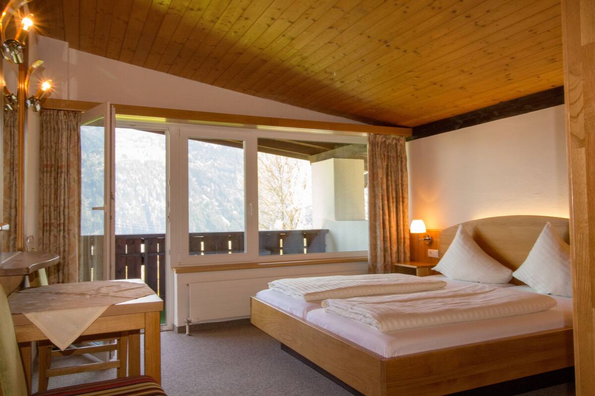 A hotel room with a wooden ceiling, a bed with white bedding, a desk with a chair, a window with curtains, and a nightstand with a lamp.