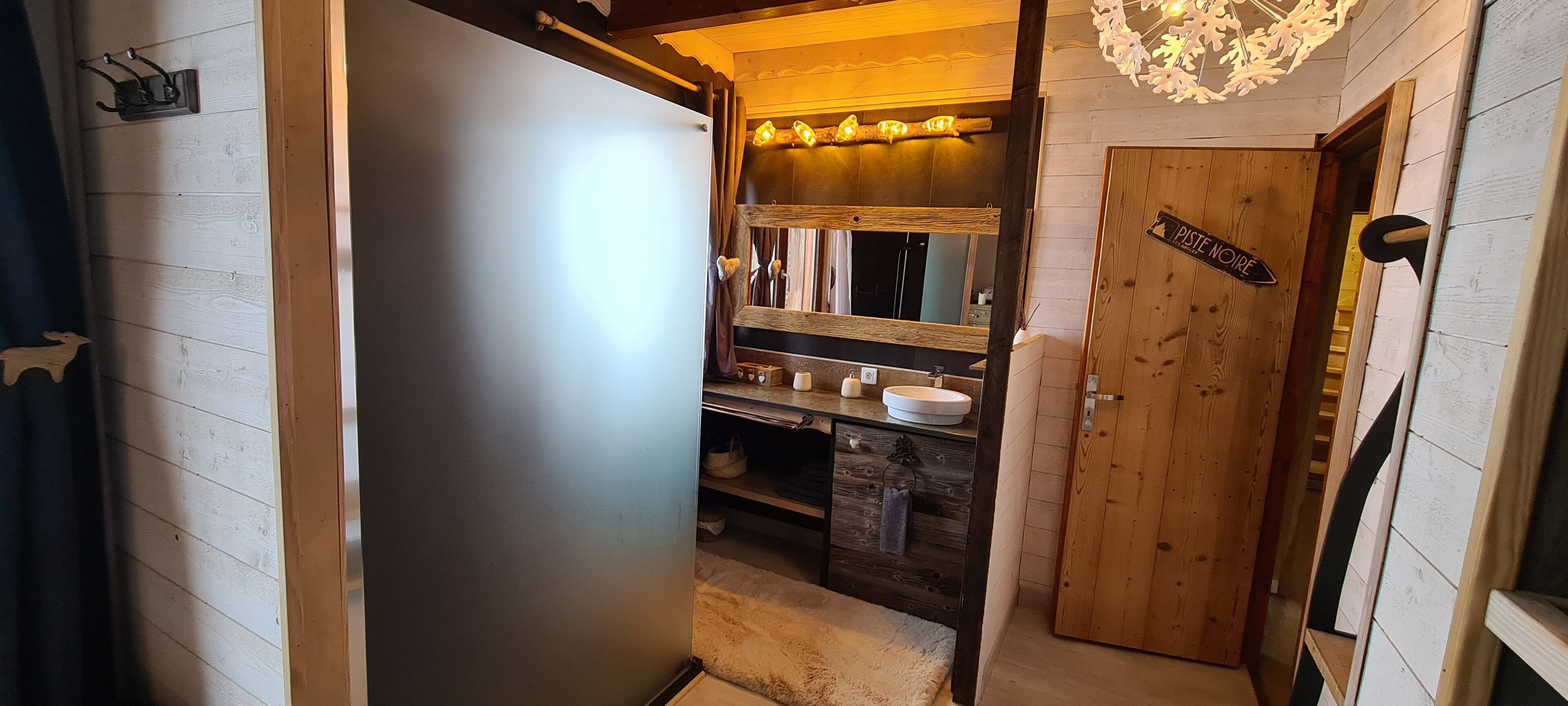 A narrow hallway with a wooden door labeled "PISTE NOIRE" leading to a bathroom with a wooden vanity, a mirror, and a chandelier.